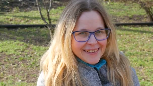 Piękna młoda kobieta z blond włosami i niebieskimi oczami w okularach siedzi w wiosennym parku i uśmiecha się - Materiał filmowy, wideo