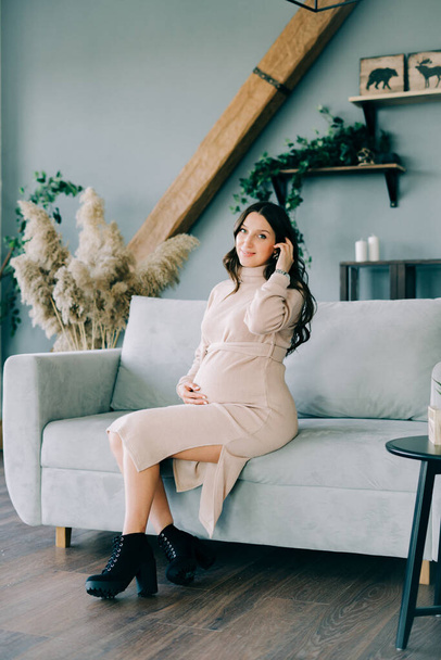 a young beautiful pregnant woman in a beige dress sitting on a gray sofa - 写真・画像