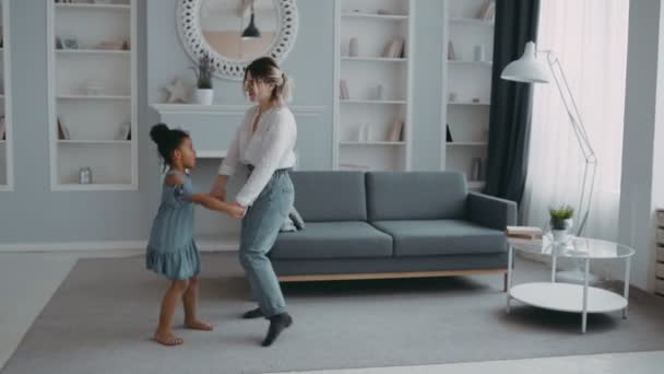Smiling active young mother nanny baby sitter holding hands with happy little kid girl, having fun together in living room. Joyful mommy dancing jumping with cute small daughter at home. - Imágenes, Vídeo