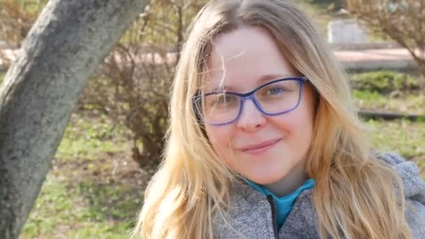 Piękna młoda kobieta z blond włosami i niebieskimi oczami w okularach siedzi w wiosennym parku i uśmiecha się - Materiał filmowy, wideo