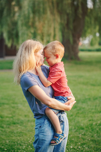 Young Caucasian mother holding boy toddler son in hands arms. Family mom kissing hugging embracing a child outdoor in park on summer day. Happy authentic family childhood lifestyle.  - Foto, Imagem