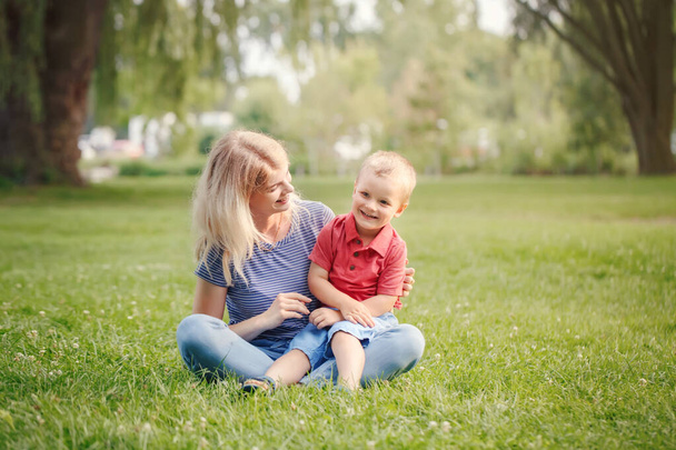 Young smiling Caucasian mother and laughing boy toddler son sitting on grass in park. Family mom and child hugging having fun outdoor on summer day. Happy authentic family childhood lifestyle.  - Фото, зображення