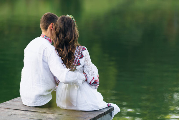 Ένα κορίτσι και ένας άντρας με κεντημένα ρούχα κάθονται σε μια ξύλινη προβλήτα και κοιτάζουν τη λίμνη. πίσω όψη. - Φωτογραφία, εικόνα