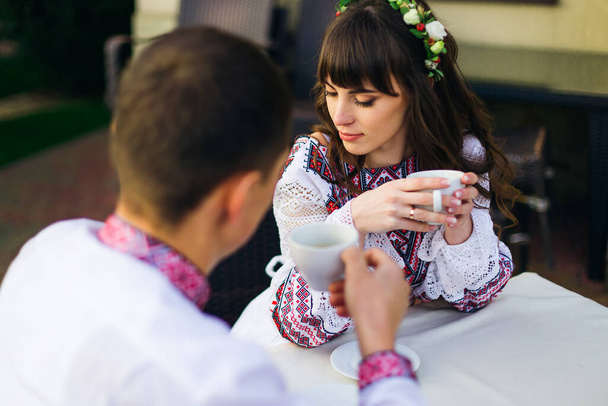 невеста закрыла глаза и держала чашку горячего напитка, а жених смотрел на нее. молодожены в вышитой одежде в кафе
. - Фото, изображение