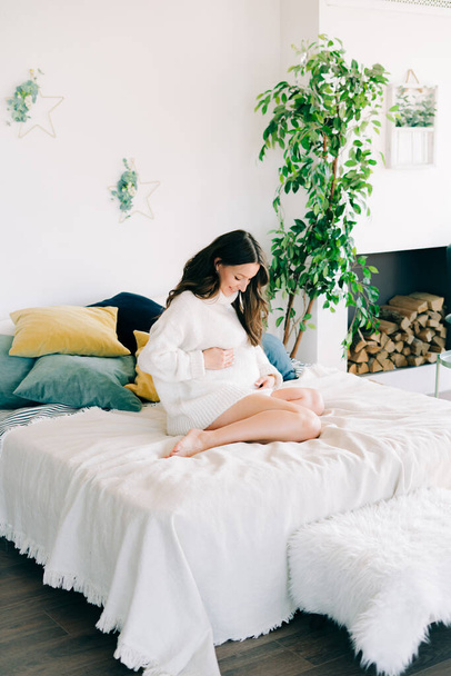 fiatal boldog terhes nő fehér meleg pulóverben ül az ágyon, és tartja a gyomrát - Fotó, kép