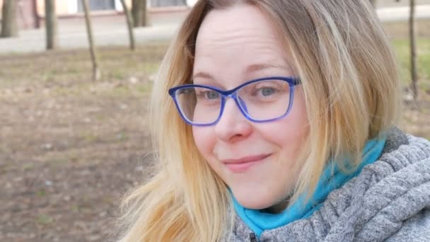 Πορτρέτο μιας όμορφης νεαρής γυναίκας με ξανθά μαλλιά και μπλε μάτια που φοράει γυαλιά κάθεται σε ένα πάρκο της άνοιξης και χαμογελά και κάνει πρόσωπα - Πλάνα, βίντεο