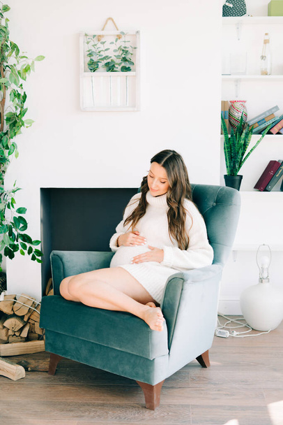 a young happy pregnant woman in a white warm sweater is sitting in a blue chair - Foto, imagen