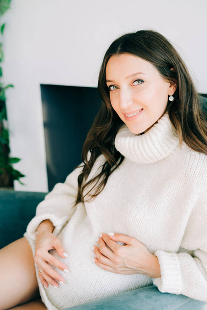 egy fiatal, boldog terhes nő fehér meleg pulóverben ül egy kék székben. - Fotó, kép