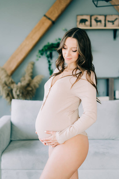 portrait of a young happy pregnant woman in a beige bodysuit is standing in a cozy spacious apartment, smiling and holding her stomach - Fotó, kép
