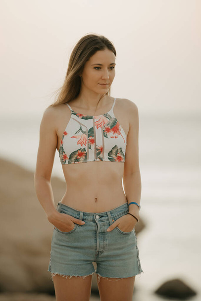 An attractive girl with long hair staying on the rocky shore of the red sea in a swim top and denim shorts on the sunrise - 写真・画像