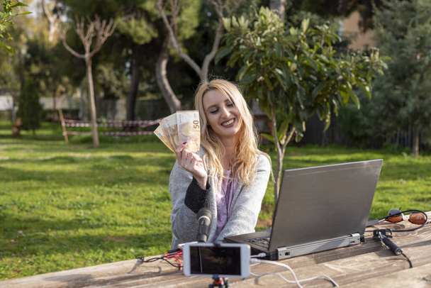 Female blond blogger demonstrates the money earned from internet services - working outdoors -  cash for review products and services - you tubers and influencers work concept - Photo, image