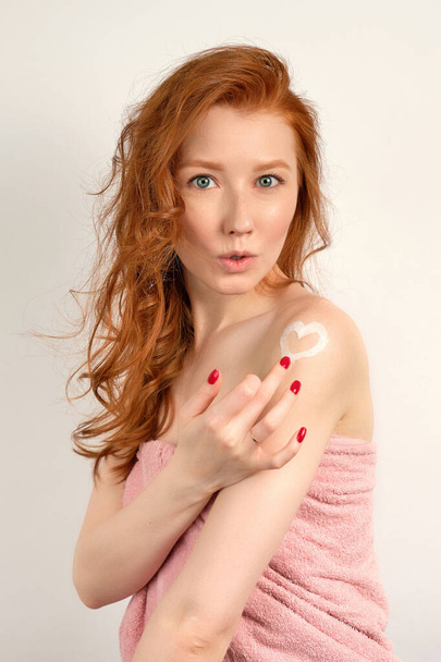 Red-haired girl with clean skin in a towel, draws a heart from a cream on her shoulder with her finger, lips extended - Fotó, kép