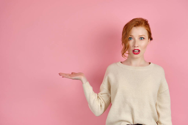 A beautiful red-haired girl with red lips in a white sweater stands on a pink background and shows palm in surprise to the side - Fotografie, Obrázek