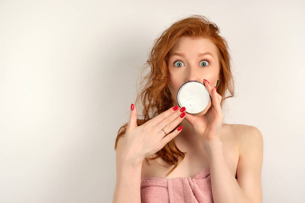 Redhead girl with clean skin looking at the camera with eyes wide open and covering mouth with a jar of cream, putting hand to her - Foto, Imagen