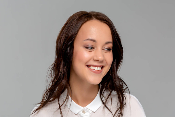 Hymyilevä nuori nainen valkoisessa paidassa poseeraa harmaan seinän taustalla studio muotokuva. Saavutus uran vauraus liiketoiminnan käsite. Mallintaa kopiotilaa
. - Valokuva, kuva