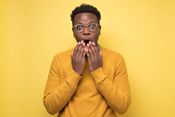 Jovem afro-americano em suéter amarelo em choque, mantendo a boca bem aberta
 - Foto, Imagem