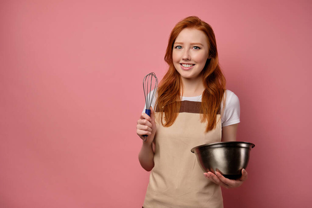 The red-haired girl in an apron stands on a pink background and smiles, holding a bowl and a whisk in her hands. - Foto, immagini