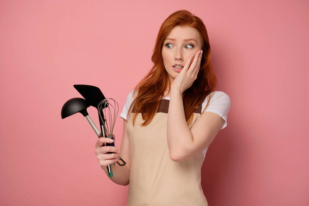 A redhead girl in an apron stands on a pink background with a shovel, a whisk and a ladle in her hand and looks at them in horror - Foto, afbeelding