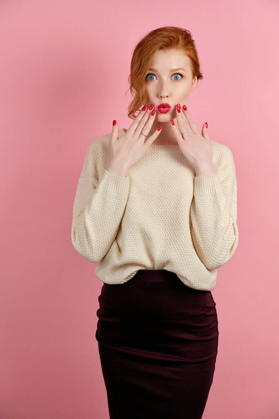 A beautiful red-haired girl with red lips and manicure stands on a pink background and looks in surprise, raising hands to face - Foto, Imagem