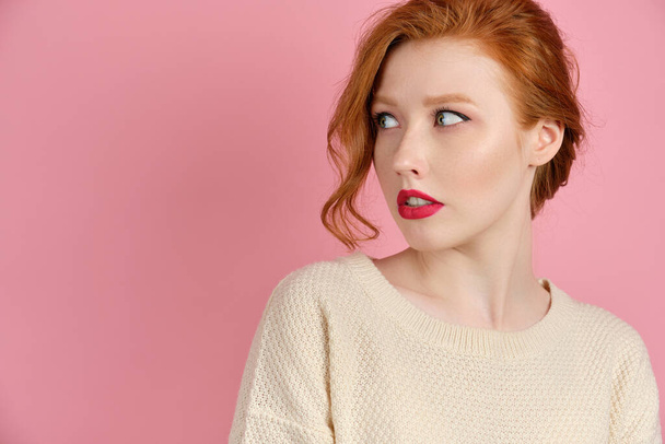 A beautiful red-haired girl with red lips in a white sweater stands on a pink background and, turning her head, looks to the side - Foto, Bild