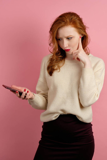 The red-haired girl stands on a pink background in a half-turn and frowns with displeasure at the phone in her hand. - Foto, Bild