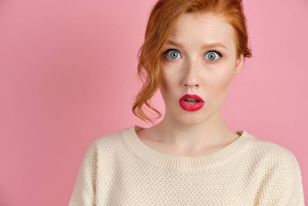Beautiful red-haired girl with red lips in a white sweater stands on a pink background in surprise opening her eyes and mouth - Photo, image