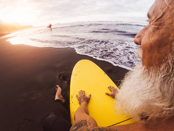 Happy surfer senior siedzi na desce surfingowej oglądając zachód słońca - Dojrzały brodaty człowiek bawiący się w dniu surfingu - Extreme sport i zdrowie ludzie koncepcja stylu życia - Zdjęcie, obraz