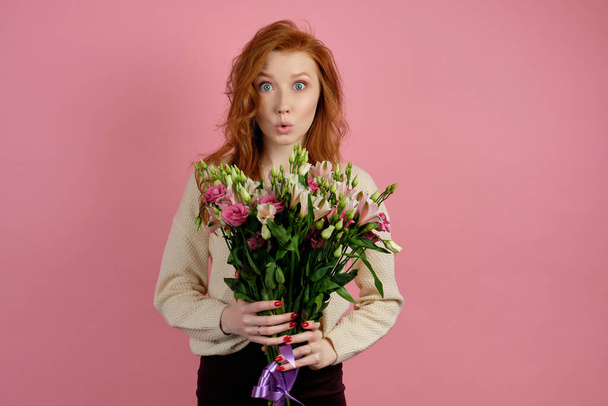 Beautiful red-haired girl holding a bouquet while standing on a pink background and opening her eyes wide and lips extended - Фото, изображение