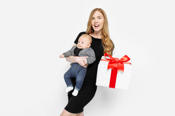 Boldog család, fiatal és gyönyörű anya gyengéden fogja a kisbabáját, kezében egy nagy ajándék doboz piros szalaggal, baba anyával egy elszigetelt fehér háttér - Fotó, kép