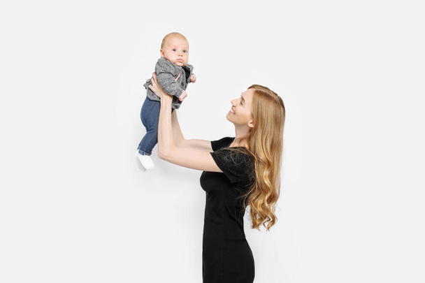 A beautiful stylish mother holds a small newborn boy in her arms and looks at him in the Studio. A beautiful fashionable woman in a black dress with long healthy hair. Isolated on a white background - Fotó, kép