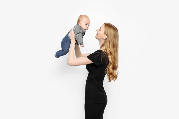 Egy gyönyörű, stílusos anya egy újszülött kisfiút tart a karjában, és ránéz a Stúdióban. Egy gyönyörű, divatos nő, fekete ruhában, hosszú, egészséges hajjal. Fehér alapon elszigetelve - Fotó, kép