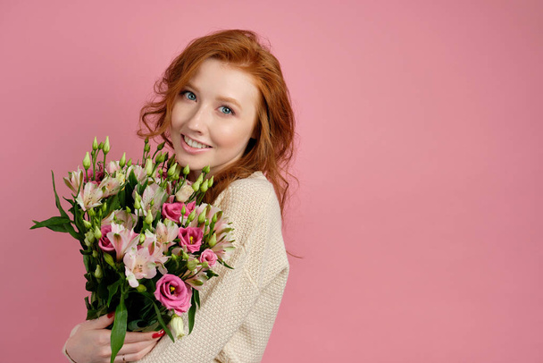 Beautiful red-haired girl hugs a bouquet with flowers on a pink background and gently smiles at the camera - Фото, зображення