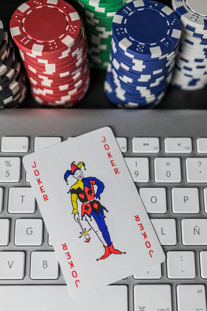 Joker karta pokerowa na klawiaturze komputera z kolorowymi kolumnami chip w tle.Widok od góry. Pojęcie przypadku, zakładów i gry online - Zdjęcie, obraz