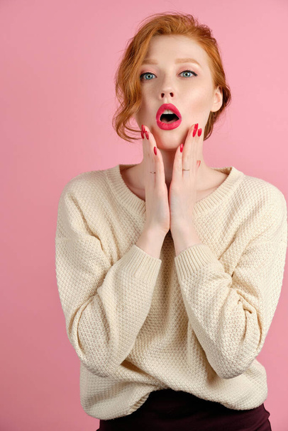 Beautiful red-haired girl with red lipstick and manicure stands on a pink background and, opening her mouth, holds hands by face - Foto, imagen