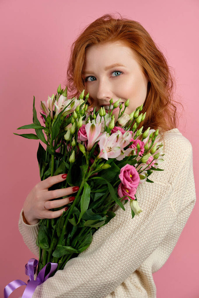 Beautiful redhead girl presses a bouquet to her face and looks cheerfully at the camera through the flowers, on a pink background - Фото, зображення