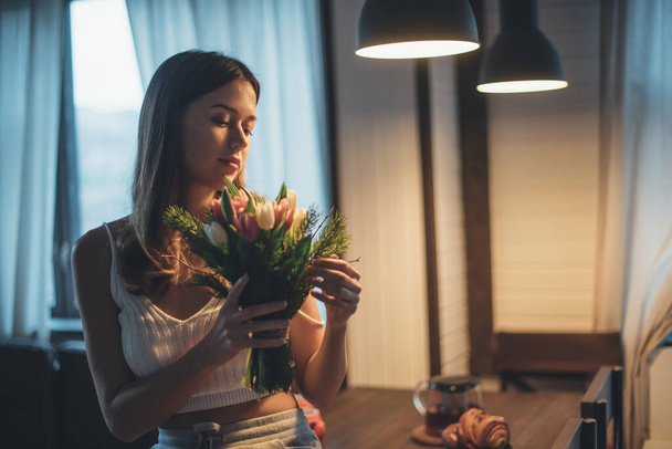 Elinde bir buket çiçekle genç bir kadının portresi. Güzel bir kız, elinde lalelerle lambaların altında bir masanın üzerinde oturuyor. Durgun ışıkta şehvetli bir portre - Fotoğraf, Görsel