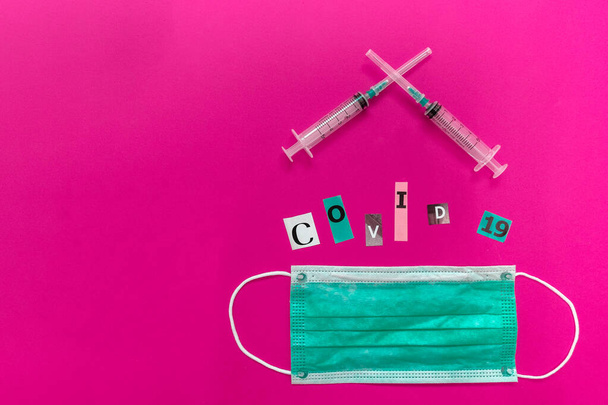 Arrangement of COVID-19 inscription, two medical syringes and surgical face mask. Letters of text are cut from a paper of magazine and put together on pink background. On the left - copy space. Flat lay - Photo, Image