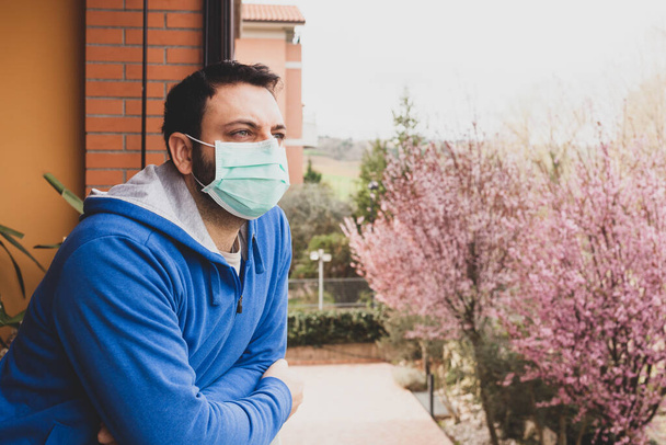 Junger kaukasischer Mann mit Maske blickt während der Quarantäne wegen Coronavirus-Pandemie auf die Hausterrasse 19. - Foto, Bild
