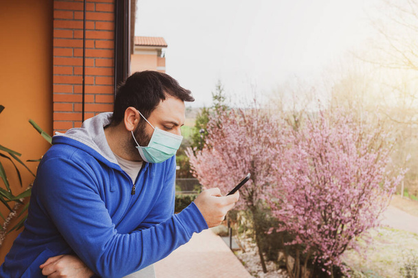 Young caucasian man with mask looking out to home terrace using mobile phone during quarantine due to coronavirus covid19 pandemic. - Photo, image