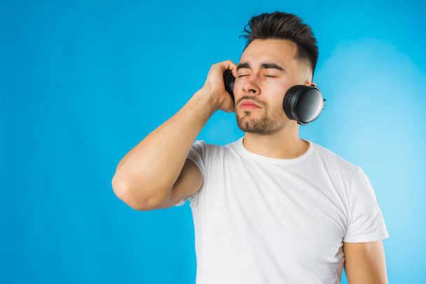 schöner europäischer junger Mann in weißem T-Shirt, der Musik hört, große schwarze Kopfhörer auf blauem Hintergrund, emotional  - Foto, Bild