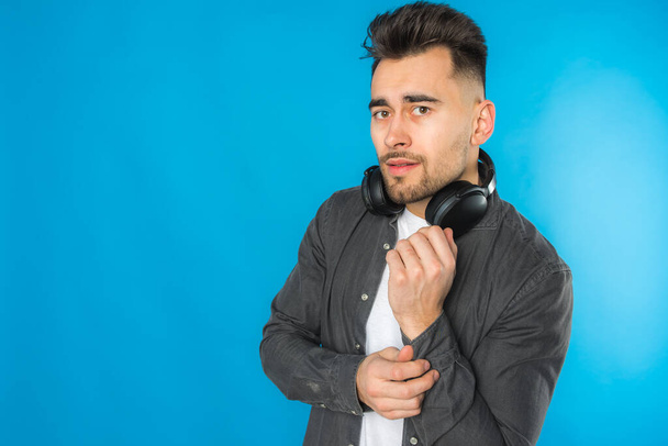 handsome european young man in gray shirt and white t-shirt listens to music, big black headphones on blue background, emotional, shows different hand gestures - Photo, Image