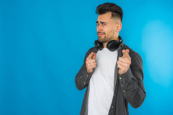 knappe Europese jongeman in grijs shirt en wit t-shirt luistert naar muziek, grote zwarte koptelefoon op blauwe achtergrond, emotioneel, toont verschillende handgebaren - Foto, afbeelding