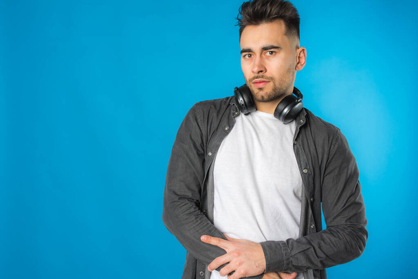 handsome european young man in gray shirt and white t-shirt listens to music, big black headphones on blue background, emotional, shows different hand gestures - Photo, Image