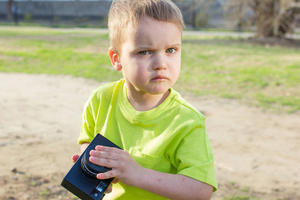 Хитрый маленький мальчик в зеленой рубашке с камерой в неверии смотрит в камеру
 - Фото, изображение