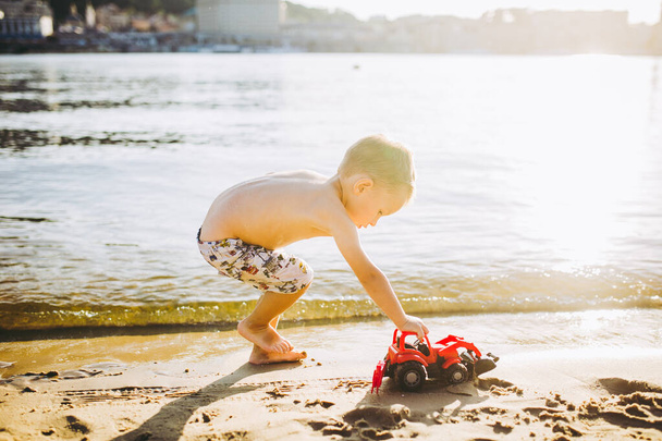 Kaukasisch kind jongen speelt speelgoed rode trekker, graafmachine op zandstrand bij de rivier in shorts bij zonsondergang dag. - Foto, afbeelding