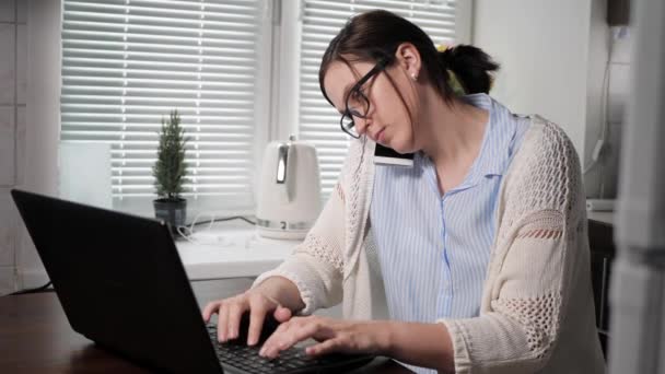 Girl freelancer at work. Attractive woman in glasses talking on phone in an uncomfortable position and at same time typing on laptop keyboard with both hands while in kitchen in apartment. Medium shot - Video