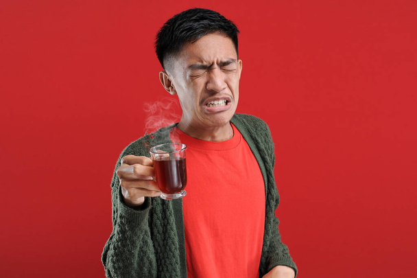 Jonge Aziatische man drinken van een kopje slechte thee / bittere thee staande over geïsoleerde rode achtergrond - Foto, afbeelding