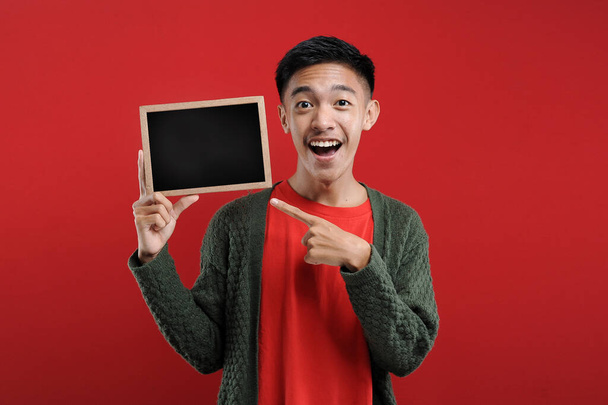 Happy excited Young Asian man holding blank small blackboard, Young Asian student smiling hold blank small blackboard, on red background - Фото, зображення