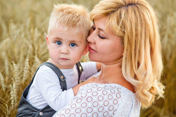 Eine schöne blonde Frau hält einen blonden Sohn auf dem Arm in einem Weizenfeld. - Foto, Bild