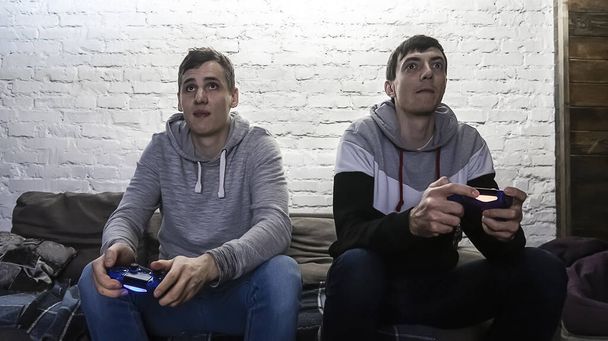 Dos hombres jugando a videojuegos frente a una pared de ladrillo blanco. Manos sosteniendo un controlador de consola. Chicos sentados en el sofá, vista frontal
 - Foto, Imagen